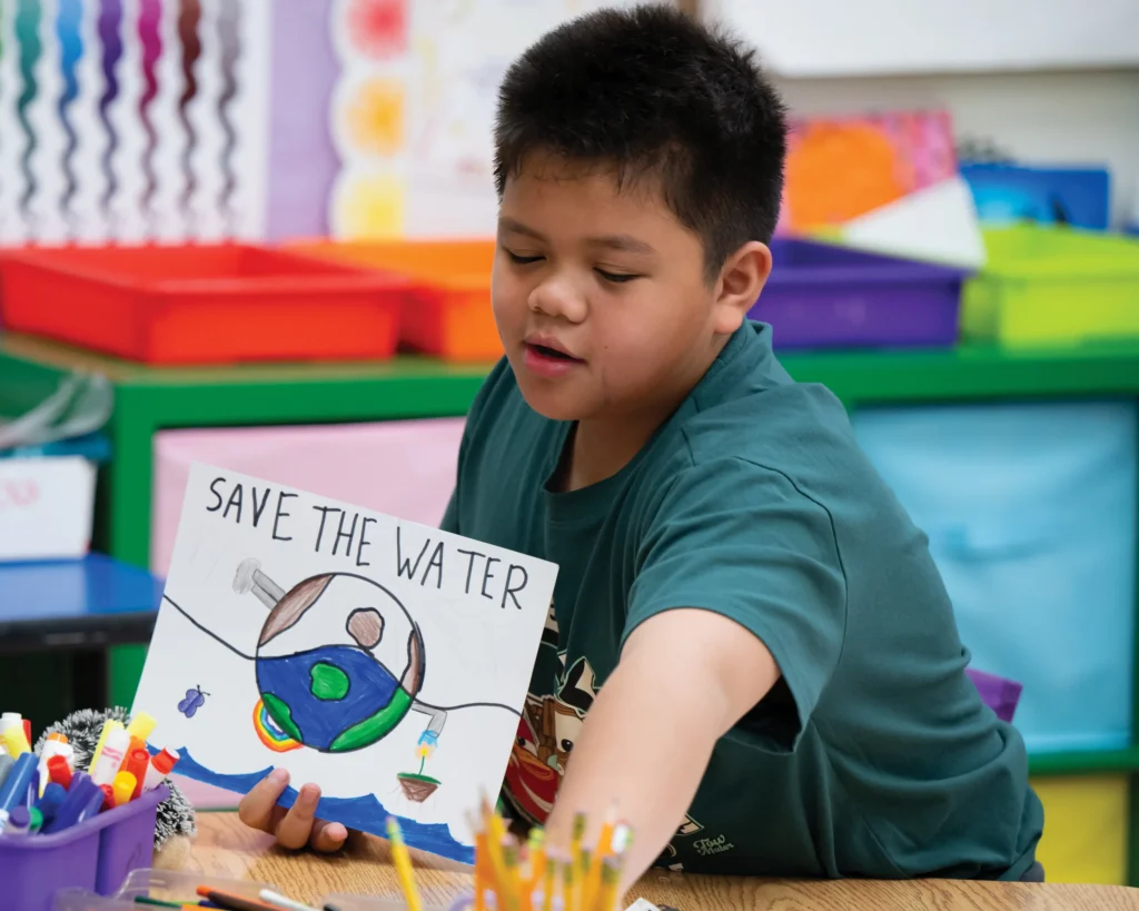 A smiling little boy at an art desk holds a picture saying: save the water