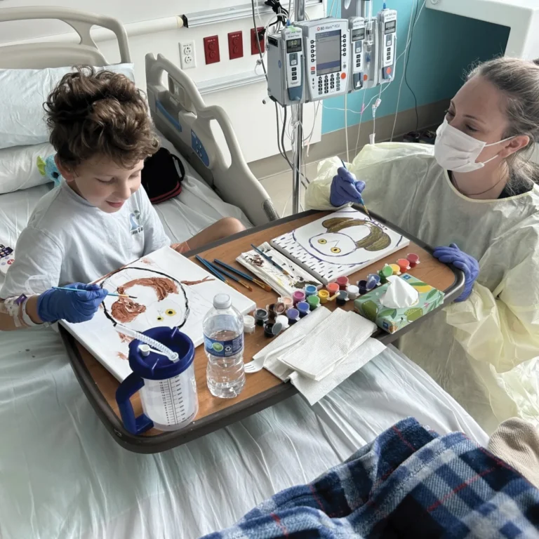 A little boy in a hospital bed paints with a masked nurse