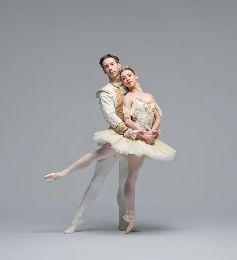 Two ballet dancers in white costumes pose against a gray background