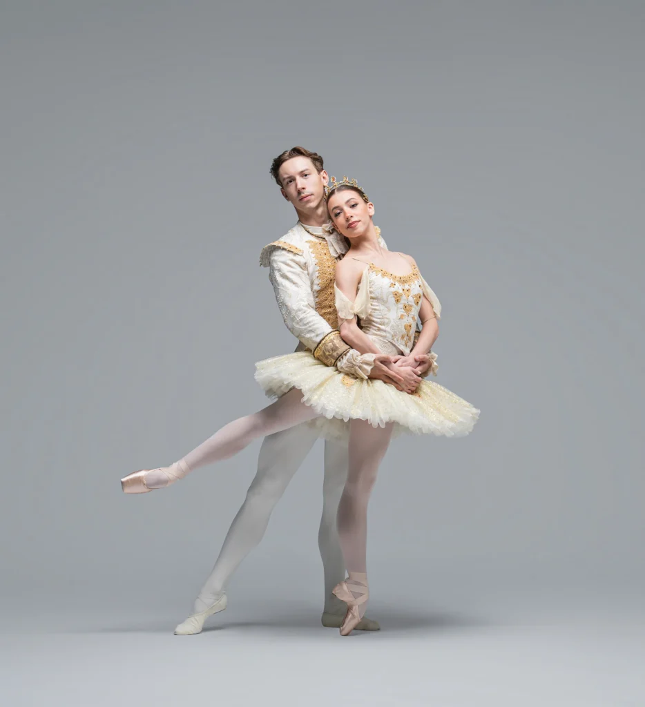 Two ballet dancers in white costumes pose against a gray background