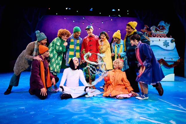 Cast of A Charlie Brown Christmas in colorful winter clothes gathered on a snowy blue stage