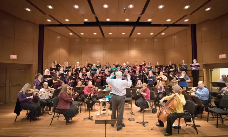 Chamber choir and orchestra performing in a wood-paneled concert hall with a conductor at center