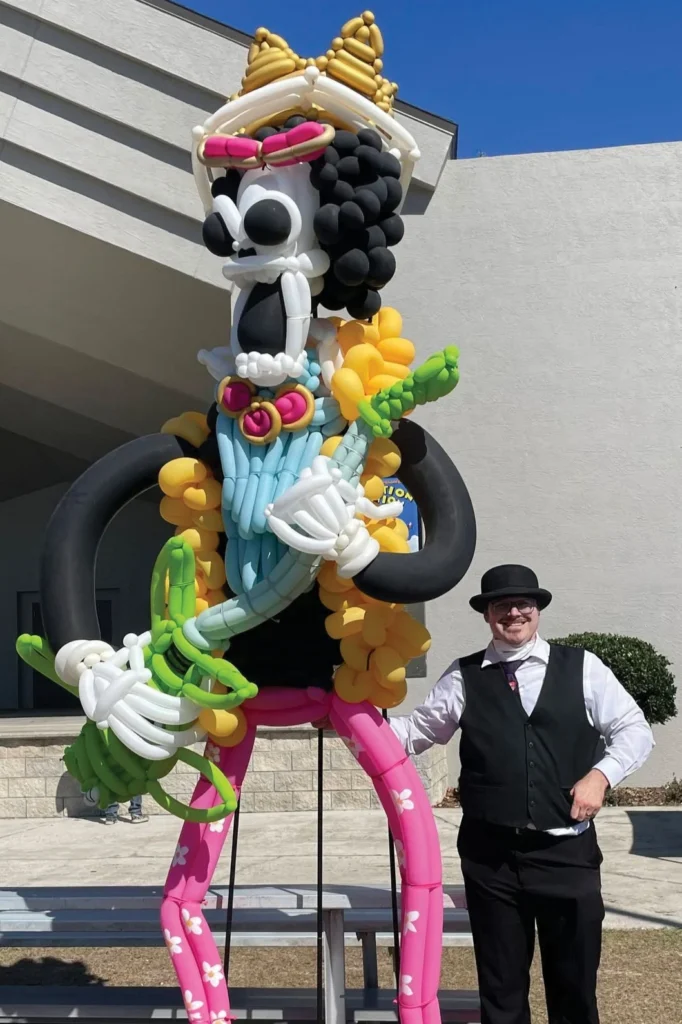 Balloon artist Bruce Carr in a top hat and vest standing beside his giant balloon sculpture of Brook from One Piece