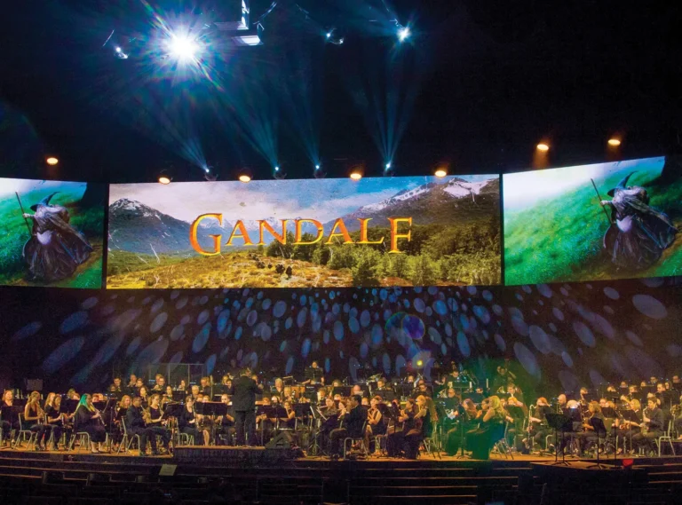 Orchestra performing under stage lights beneath large screens showing mountain scenery and the word Gandalf