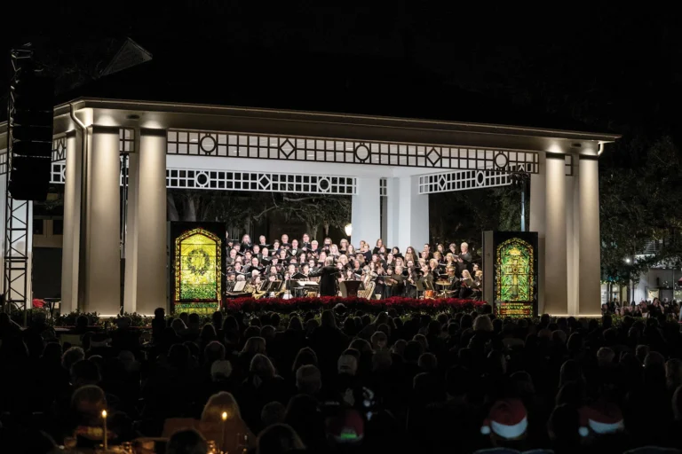 Choir performing on a lit stage flanked by illuminated Tiffany windows before a nighttime crowd in Central Park