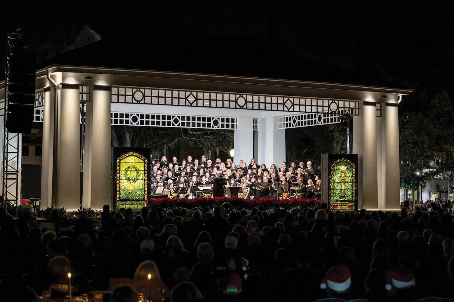 Choir performing on a lit stage flanked by illuminated Tiffany windows before a nighttime crowd in Central Park