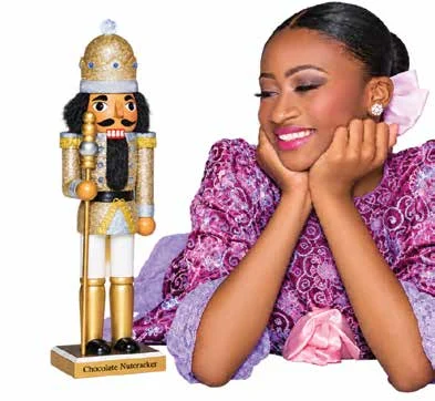 Smiling dancer in a pink and purple costume resting her chin on her hands beside a gold Chocolate Nutcracker figure