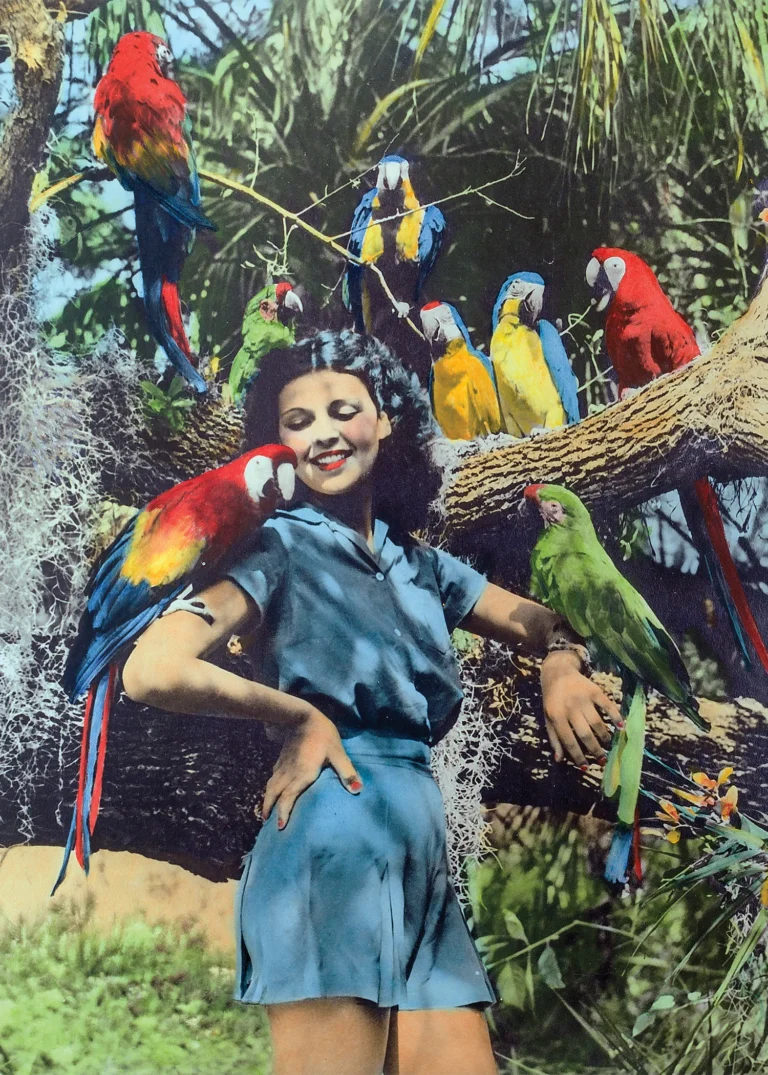Vintage hand-tinted image of a smiling woman posing with colorful macaws