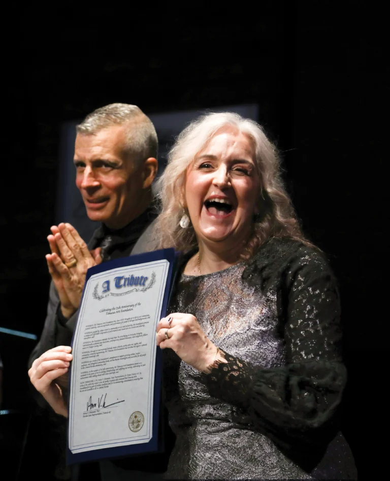 A smiling man and woman on stage, she holding a framed tribute proclamation