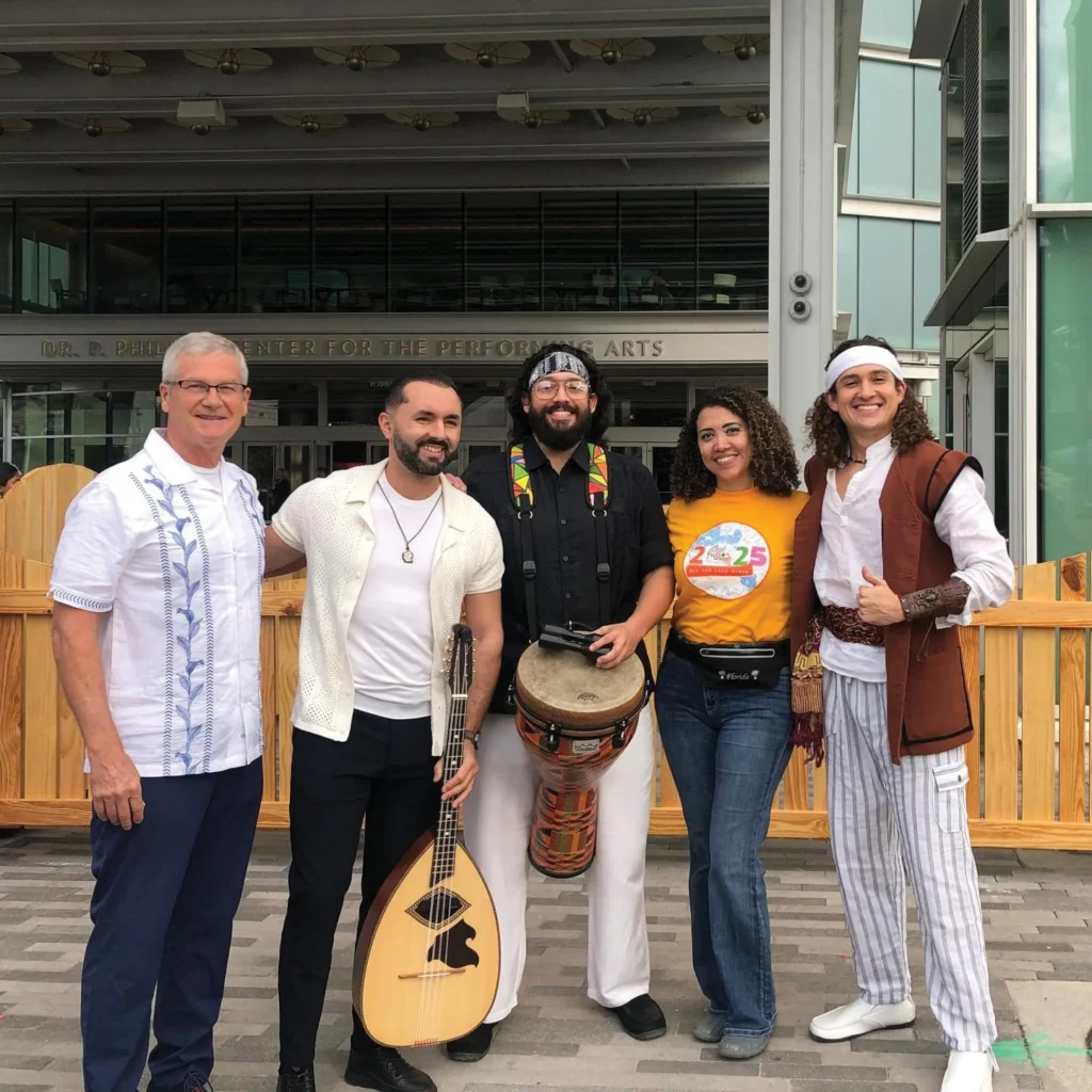 Five musicians, one holding an oud and one a hand drum, posing outdoors at FusionFest