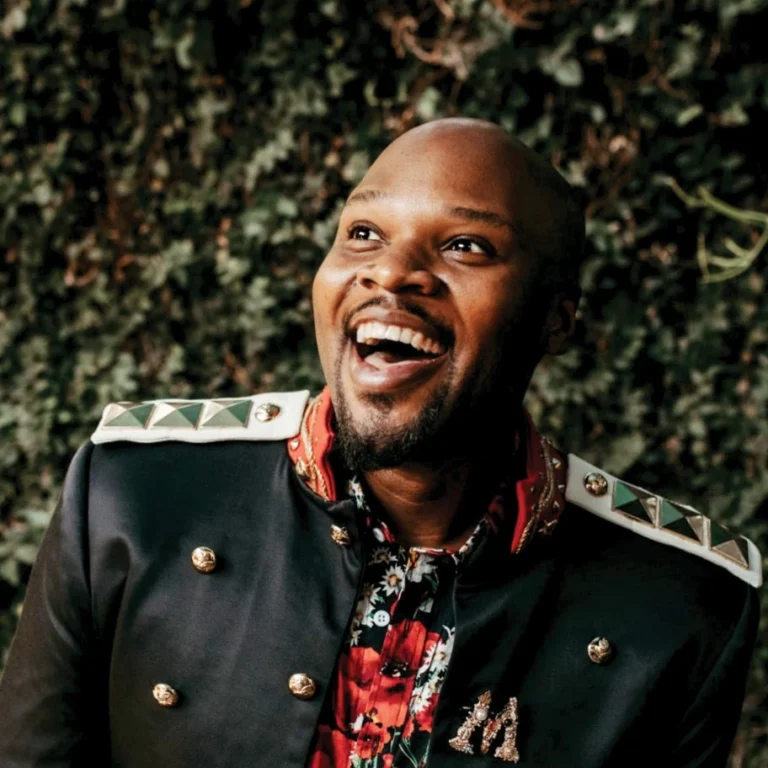 Michael James Scott laughing in a black military-style jacket and floral shirt in front of greenery
