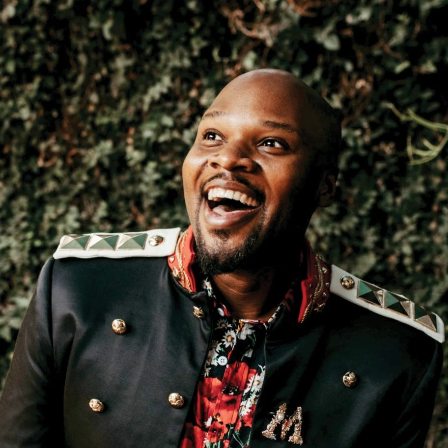 Michael James Scott laughing in a black military-style jacket and floral shirt in front of greenery