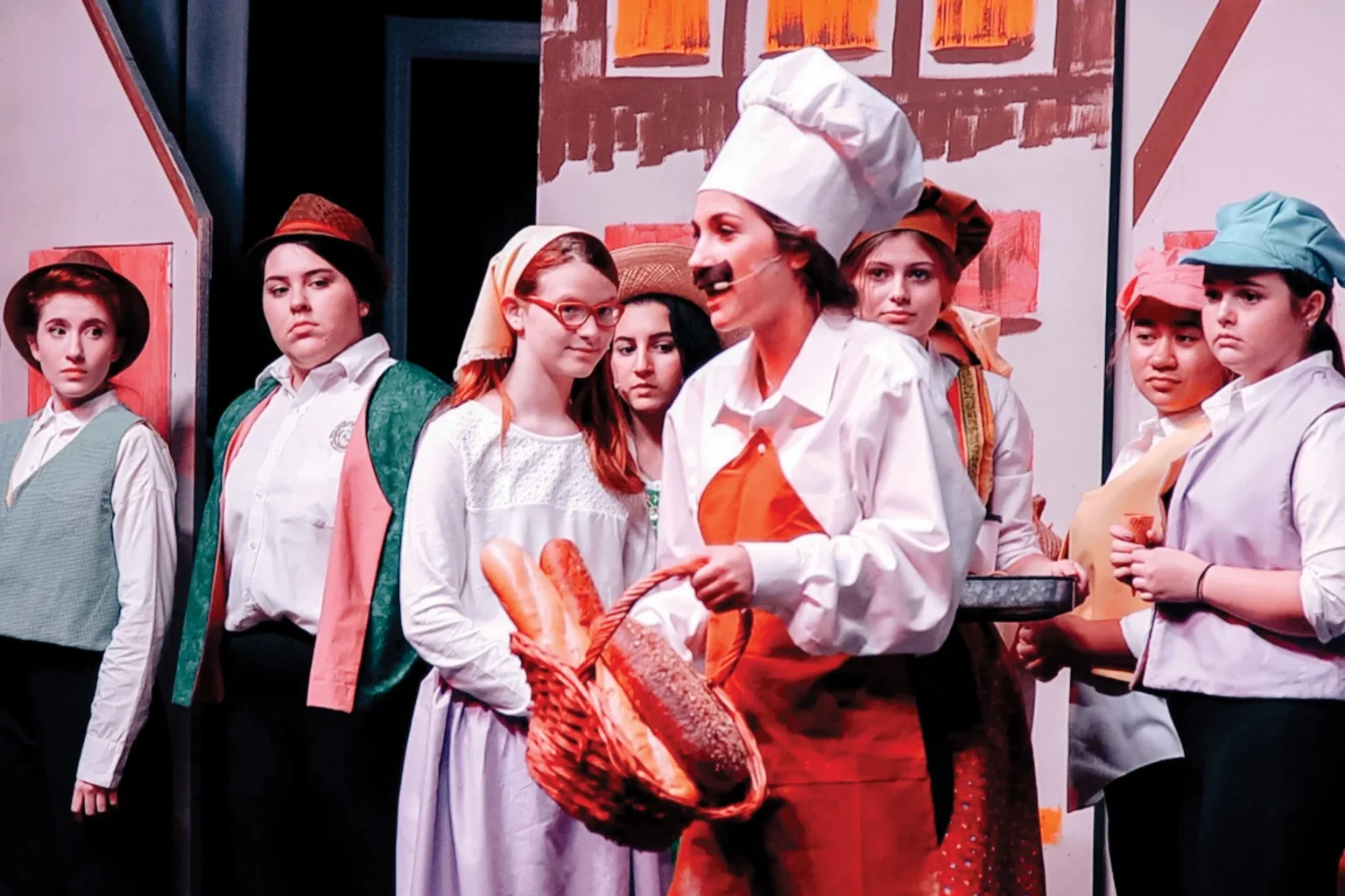 Young performers in costume on stage, including a girl in a chef’s hat holding a basket of bread