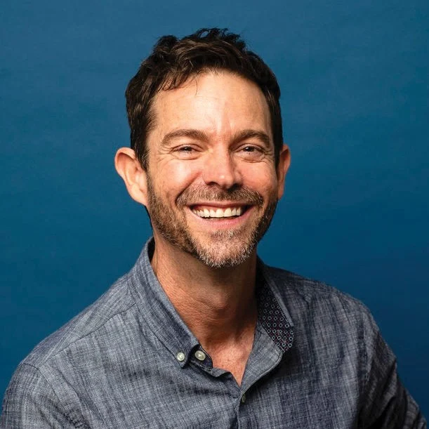 Tim Williams smiling in a gray button-up shirt against a blue backdrop